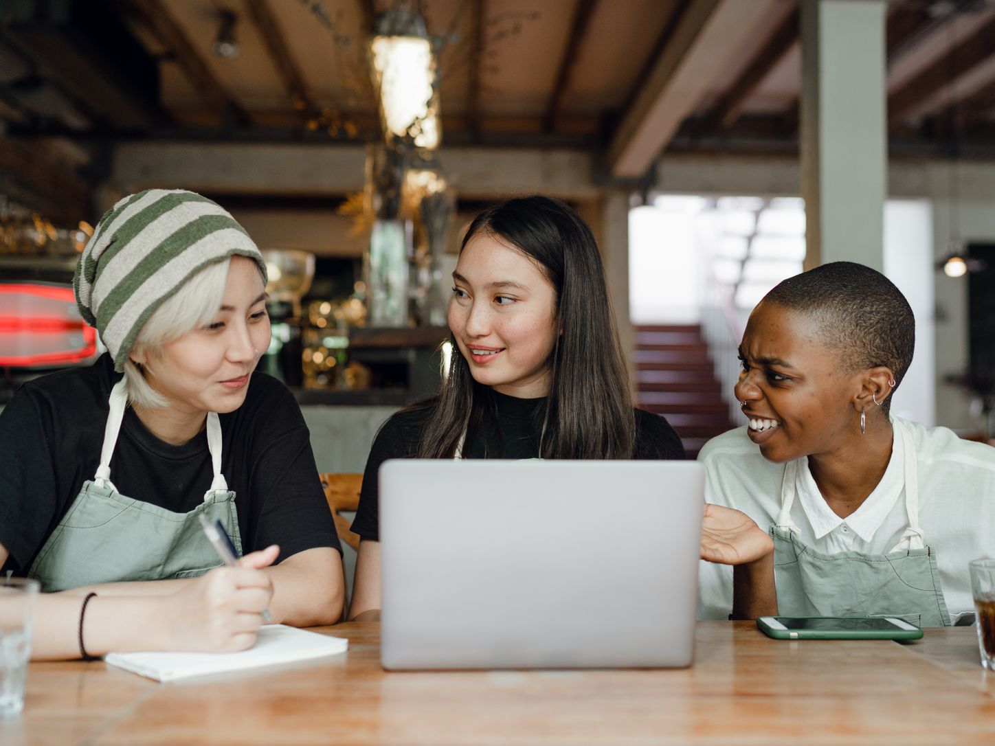 Three hospitality staff in aprons discussing work over a laptop and notebook